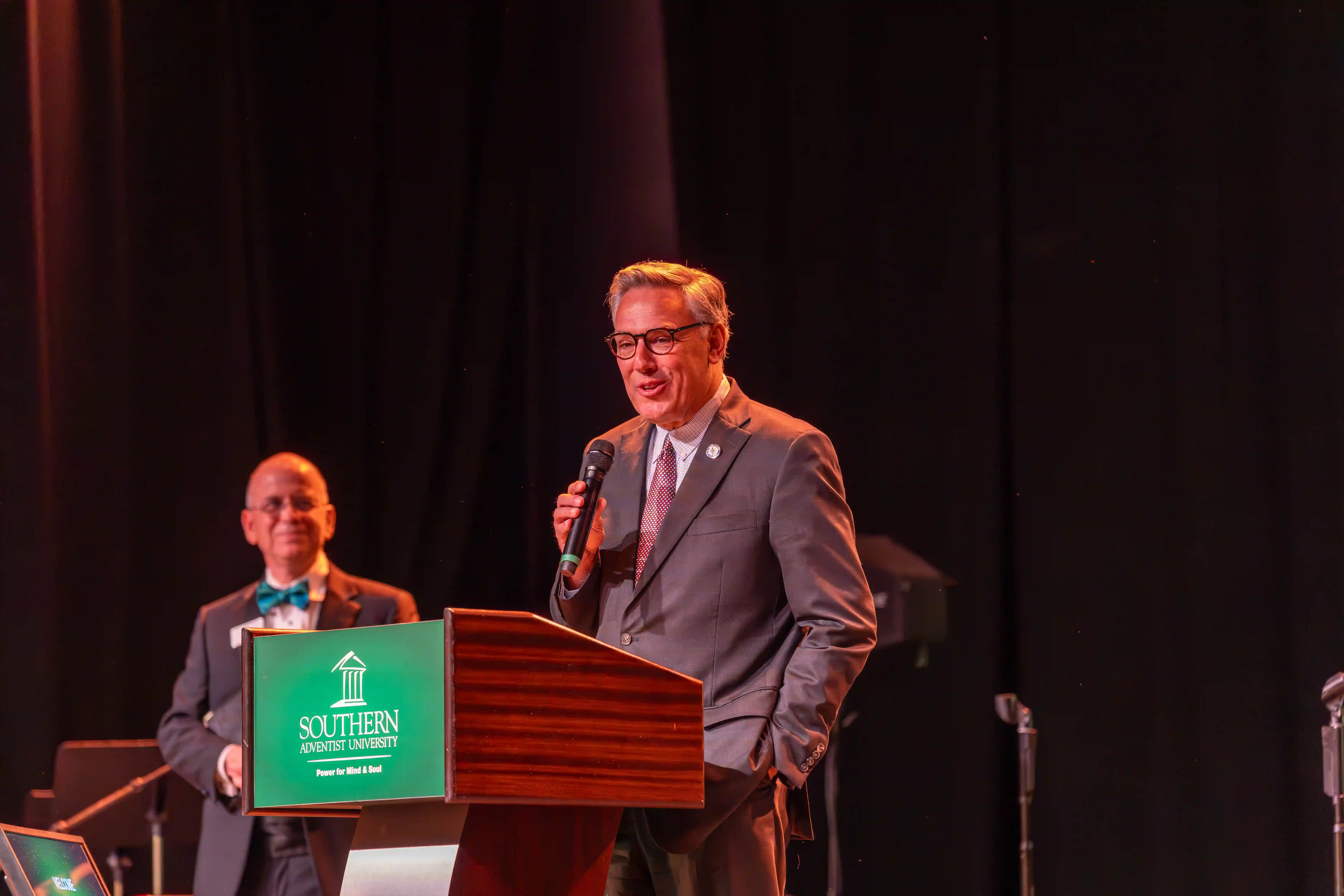 Greg Vital speaking at the SAU Podium while Ken Shaw watches from farther back on the stage